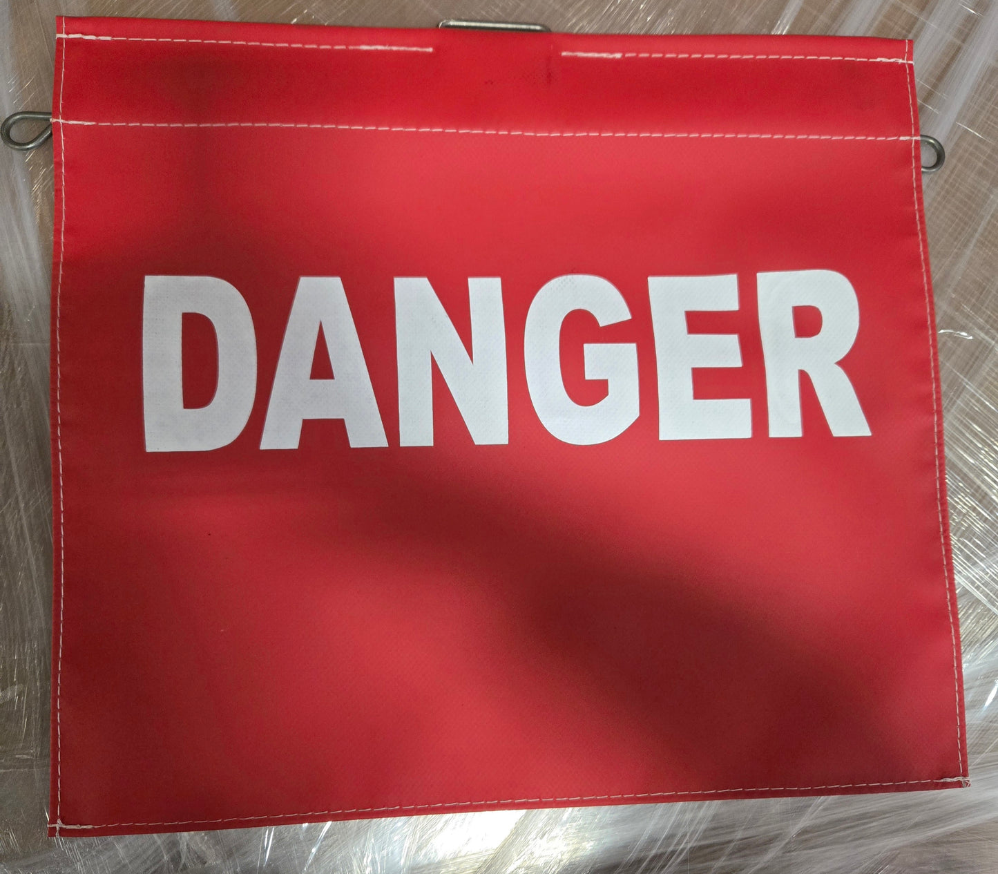 Red rectangular safety warning flag with the word “DANGER” printed in large white capital letters across the center. The flag has reinforced stitched edges and metal attachment loops visible along the sides and top. The flag is displayed on a metallic surface.