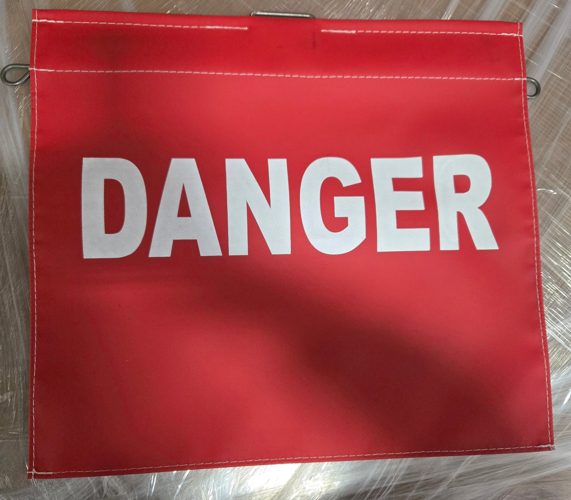 Red rectangular safety warning flag with the word “DANGER” printed in large white capital letters across the center. The flag has reinforced stitched edges and metal attachment loops visible along the sides and top. The flag is displayed on a metallic surface.