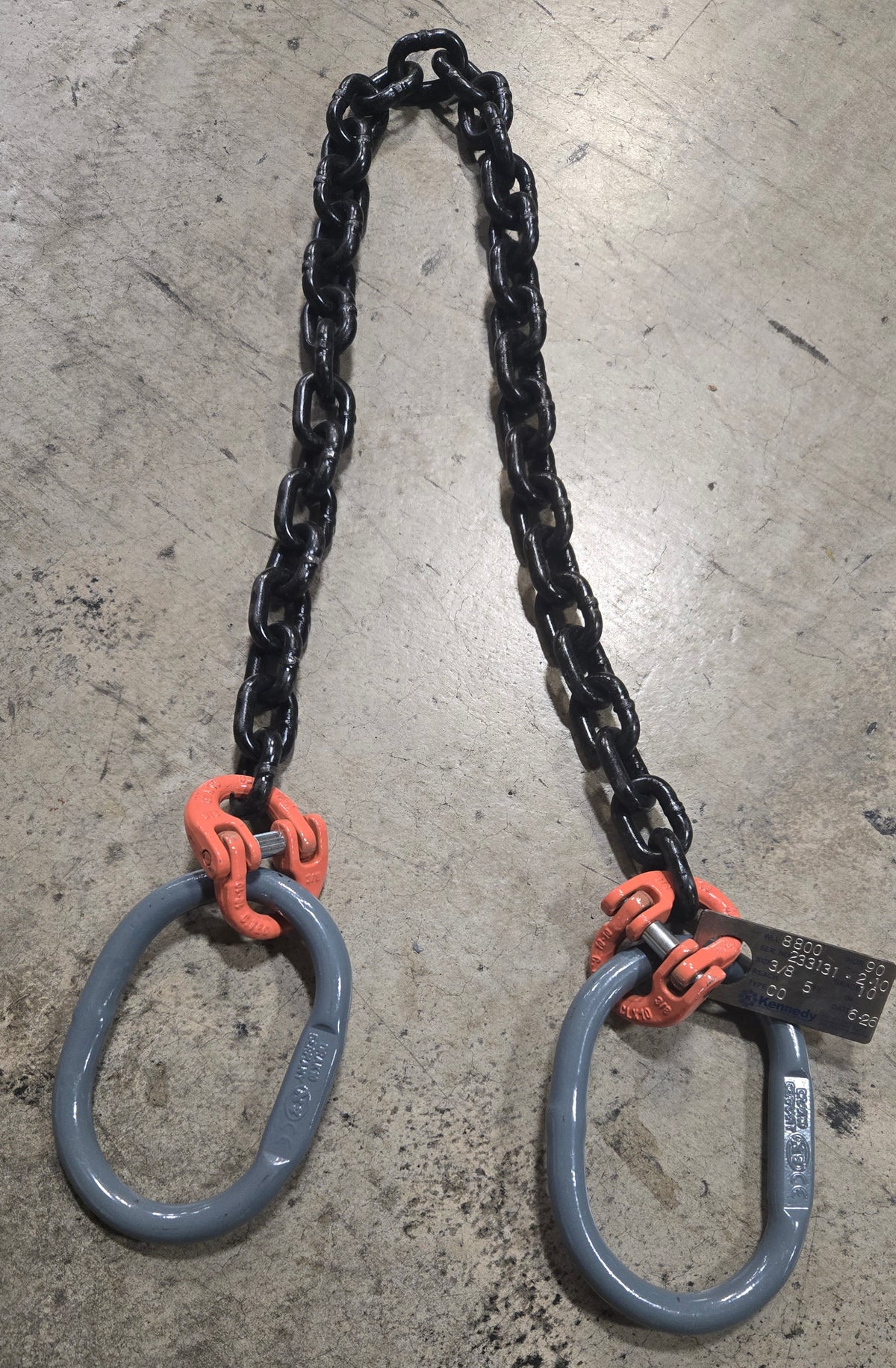 Two-leg lifting chain sling with black alloy chain legs connected to large gray oblong master links by orange coupling links. The rigging assembly is laid on a worn concrete floor, with an identification tag attached near one of the connection points.