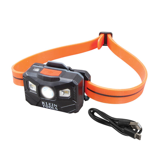 Rechargeable LED headlamp with a black lighting unit and bright orange adjustable head strap, displayed on a light gray background. The front of the headlamp features a central spotlight, two side LED lights, a power button on top, and "KLEIN TOOLS" branding on the front. A USB charging cable is shown beside the headlamp.Provide your feedback on BizChat
